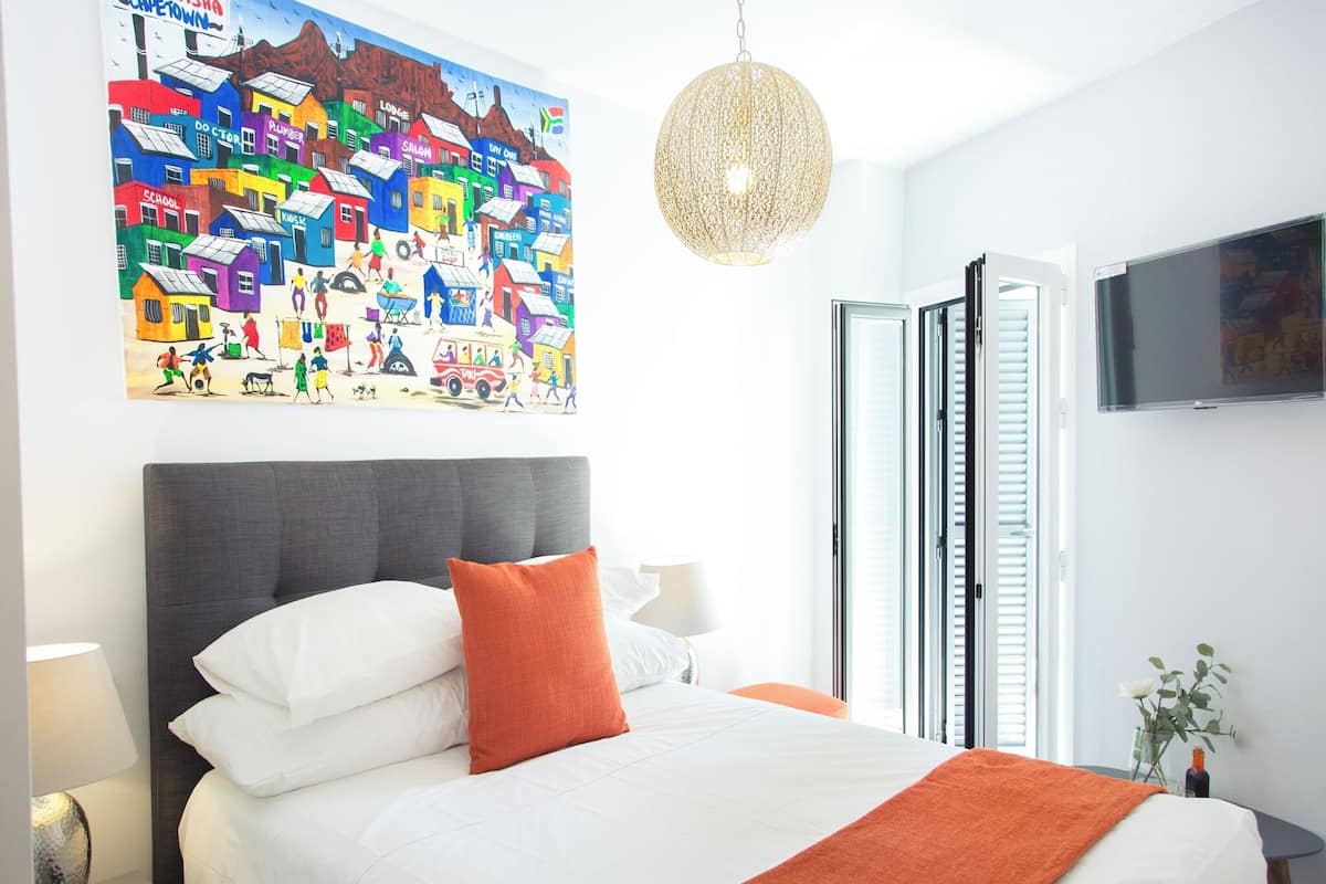 A hotel room with a bed, a large colorful artwork on the wall, a hanging light fixture, and a TV mounted on the wall.