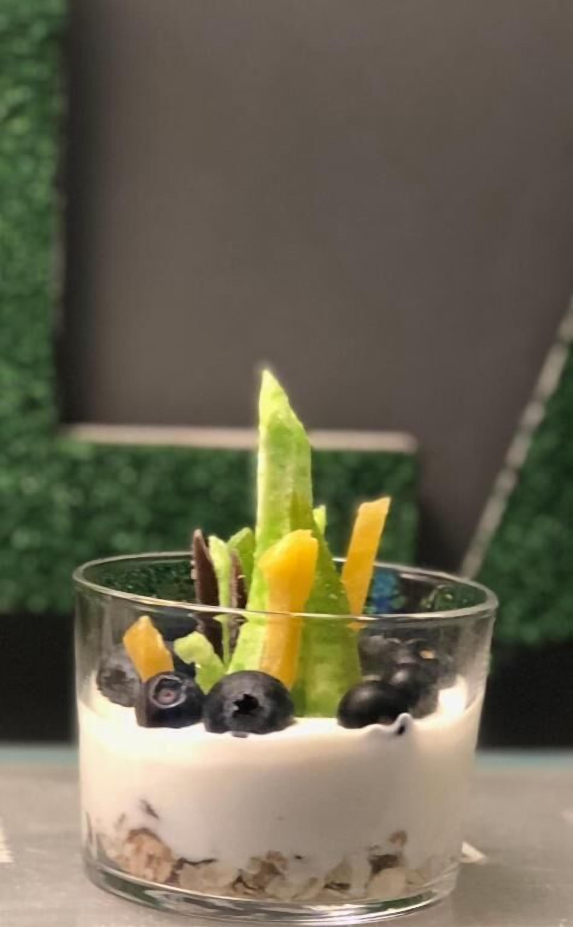 Un dessert servi dans un verre, composé de yaourt, de myrtilles, de mangue et d’un légume vert, peut-être un concombre, posé sur une table.