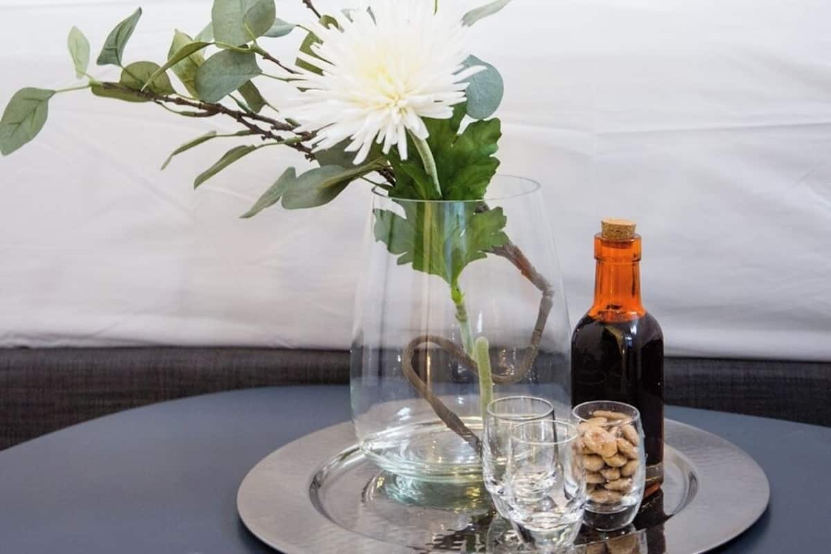 A hotel room with a bed, an orange pillow, a glass vase with flowers, a bottle, and a tray with glasses and nuts.