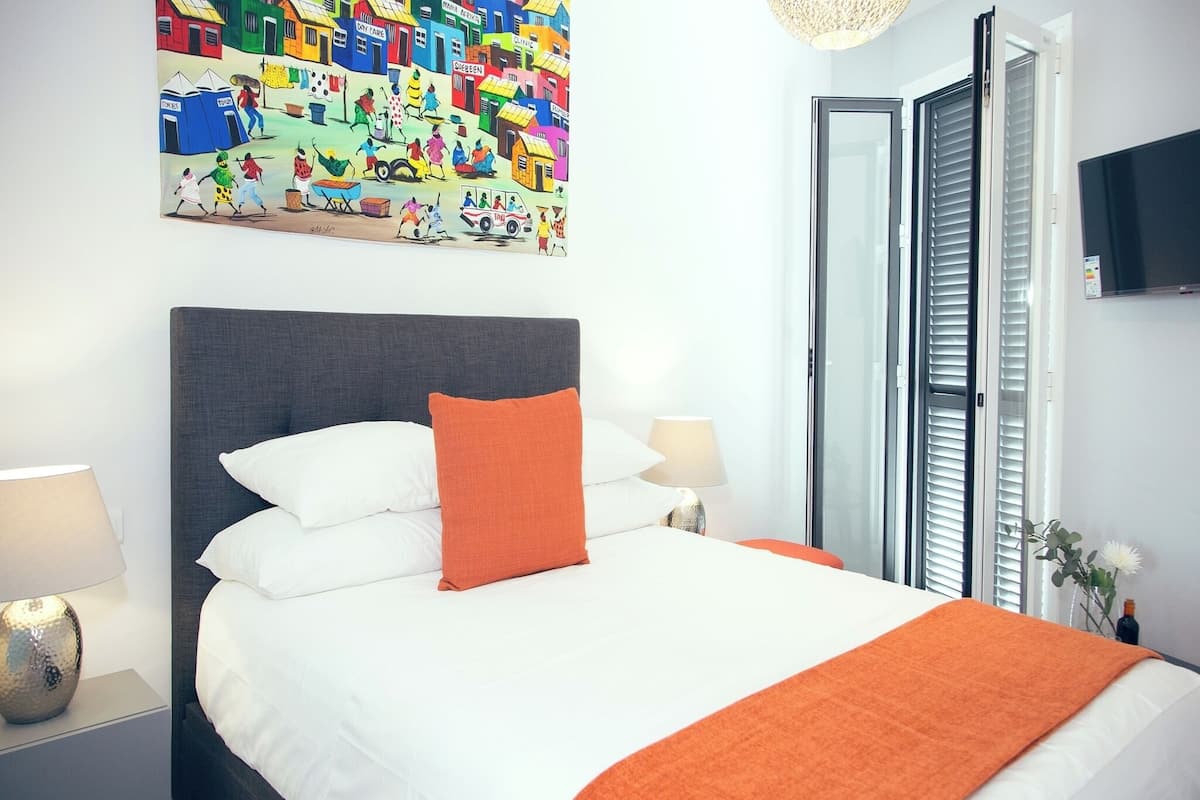 A hotel room with a large bed, two bedside tables, a nightstand, a wall-mounted TV, and a window with blinds.