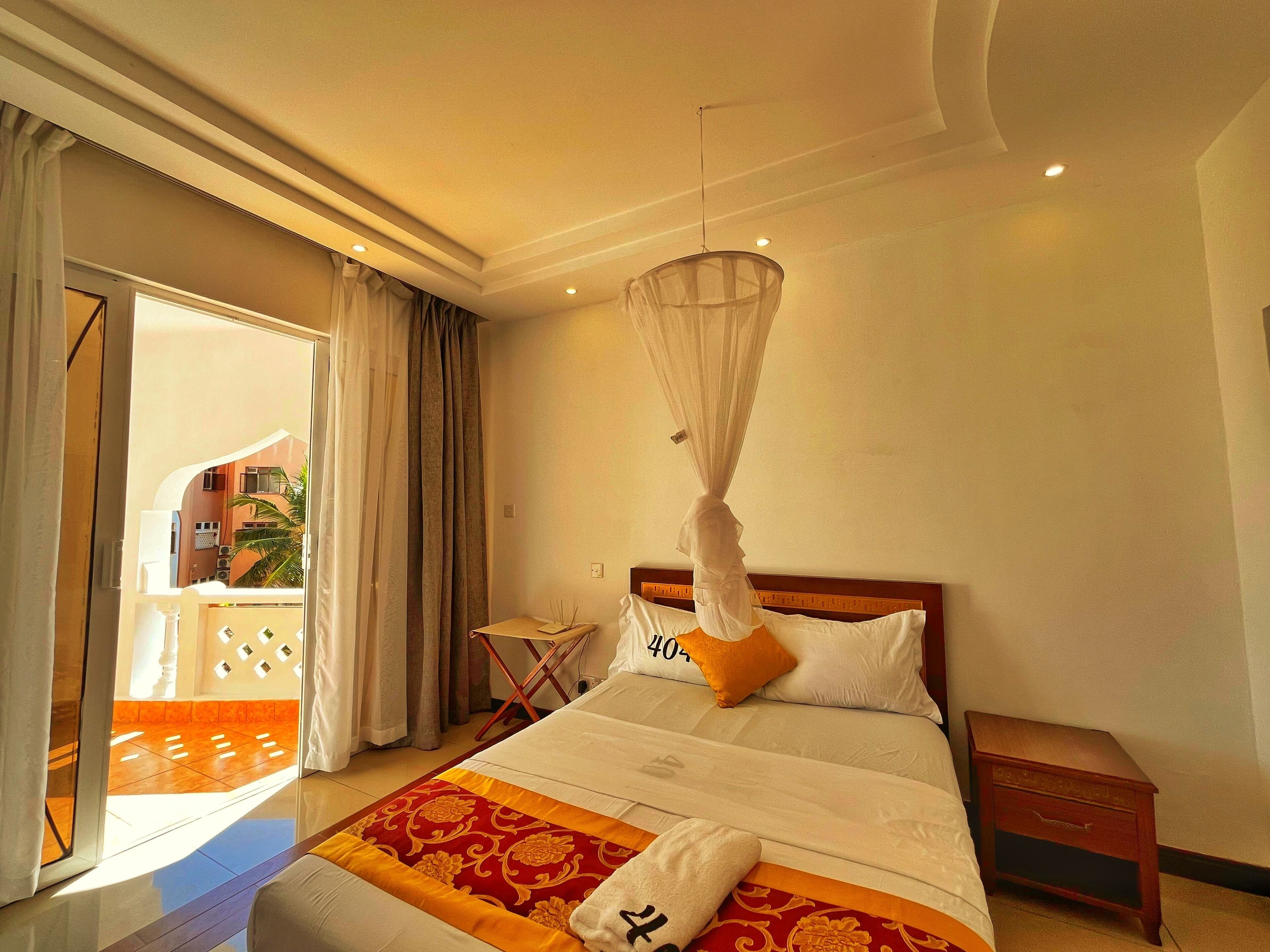 A hotel room with a bed, a bedside table, a nightstand, a window with curtains, and a view of a building outside.