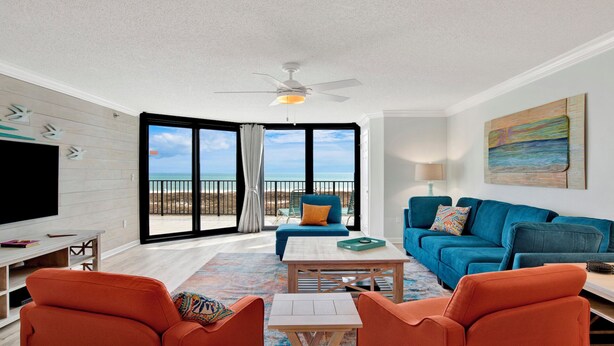 Una sala de estar con televisor, sillones naranjas, sofá azul y balcón con vista a la playa.