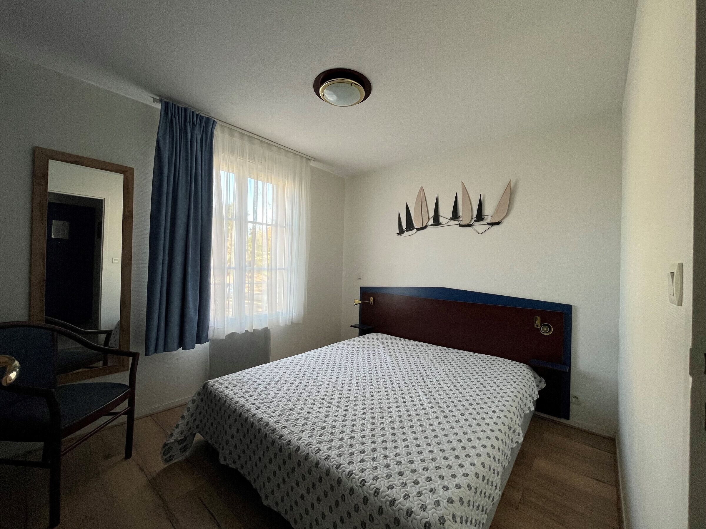 Une chambre d’hôtel avec un lit, une chaise, une fenêtre avec des rideaux, un luminaire fixé au mur et une porte.