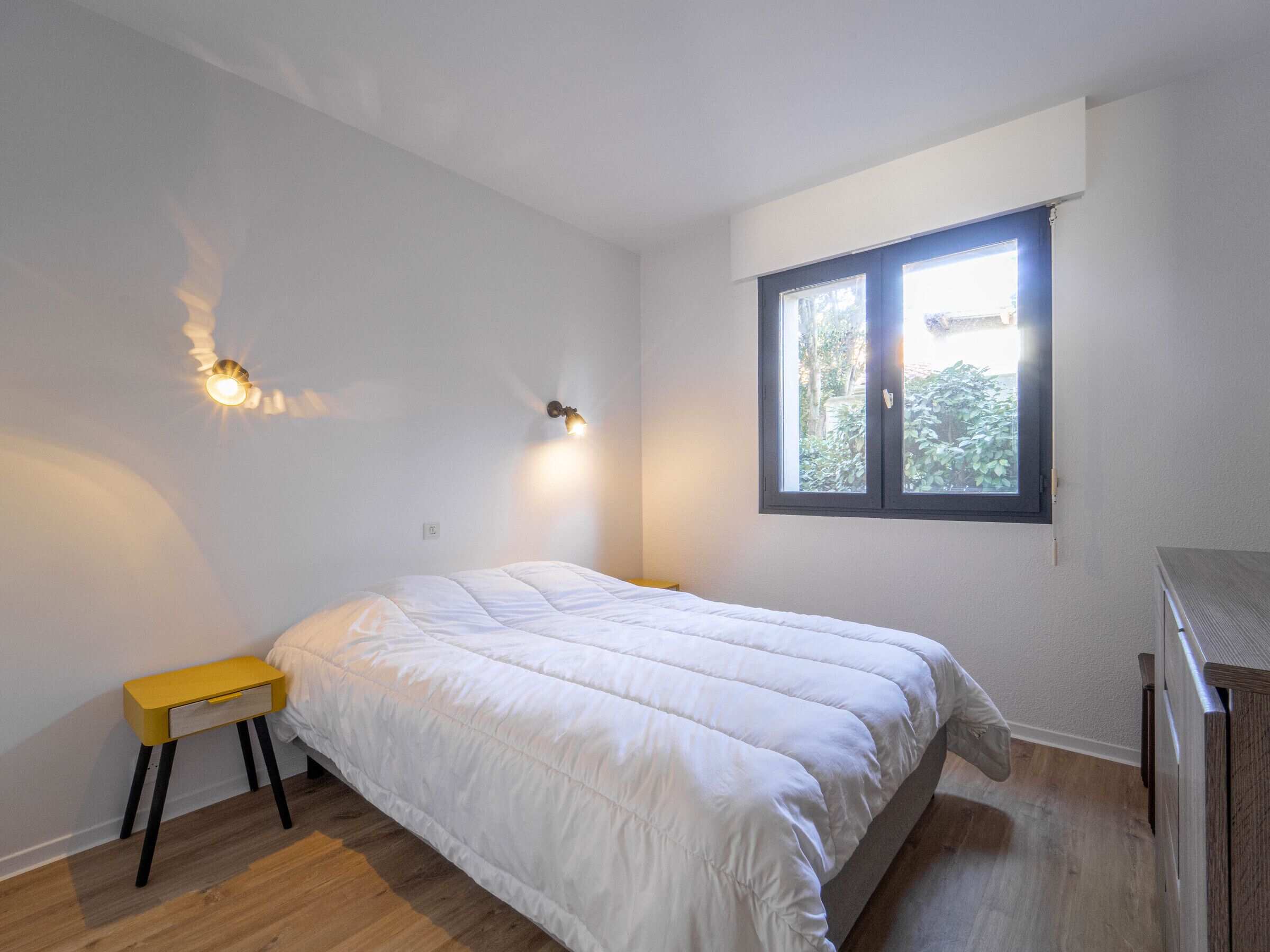 A bedroom with a bed, a small yellow bedside table, a window with a view of greenery, and a wall-mounted light fixture.