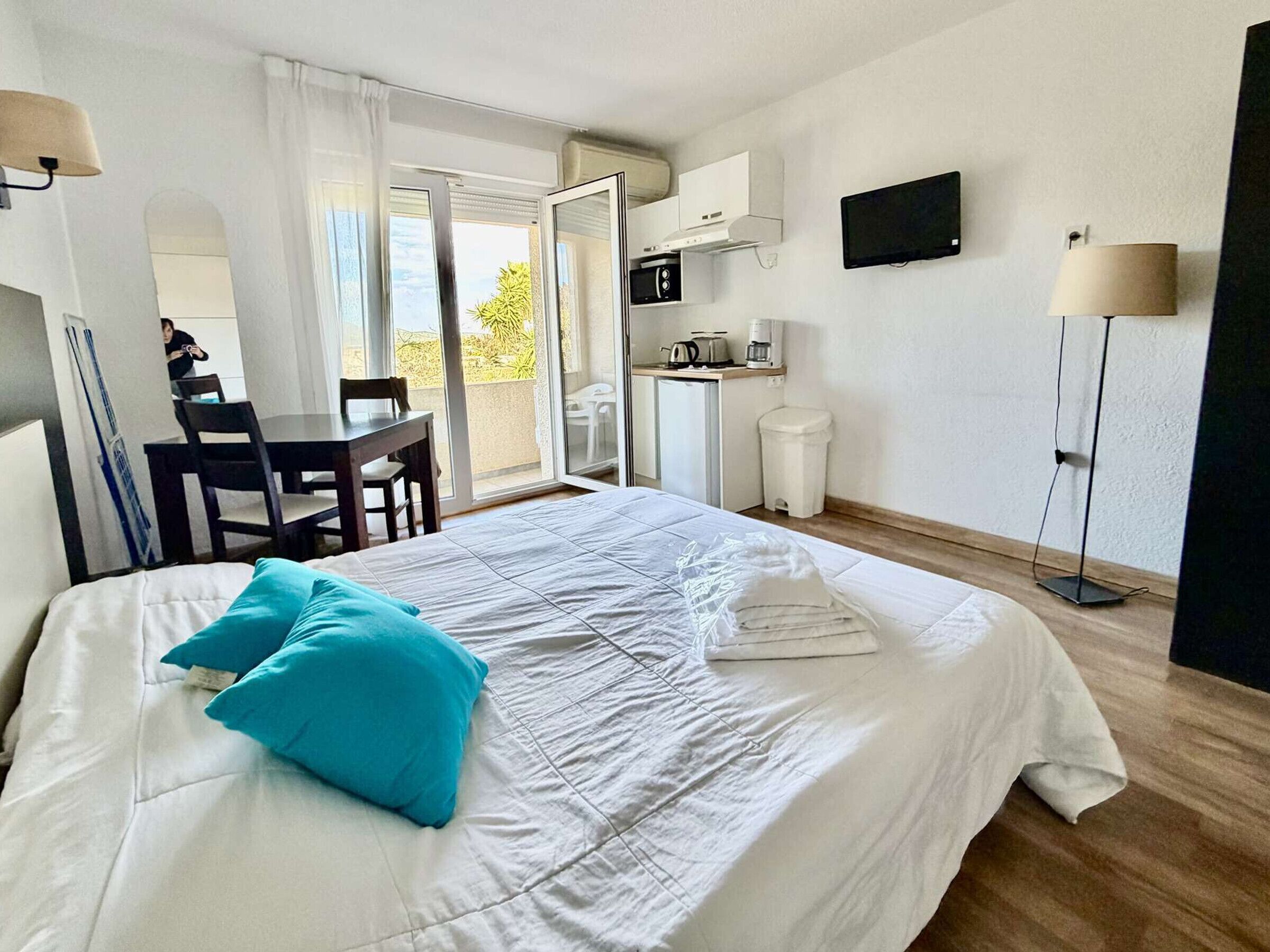 Habitació d'hotel amb llit, escriptori, cadira, petita cuina i televisió, i porta corredissa al balcó.