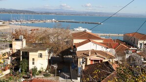 Une ville côtière aux bâtiments aux toits de tuiles rouges, un port de plaisance avec des bateaux, et une vue dégagée sur la mer et les montagnes lointaines.