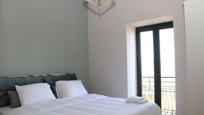 Un lit bien fait, avec du linge de lit blanc et des oreillers, un luminaire au plafond et une vue sur un balcon par une grande fenêtre.