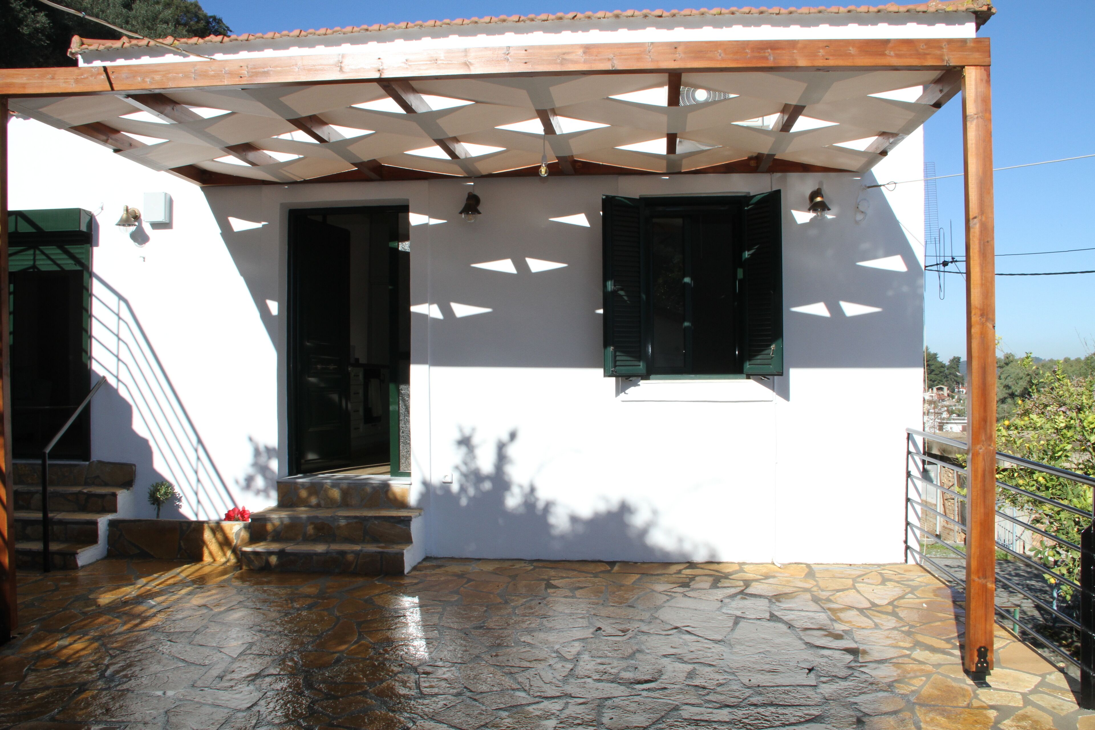 A white building with a wooden pergola, stone flooring, and a staircase leading to a door.
