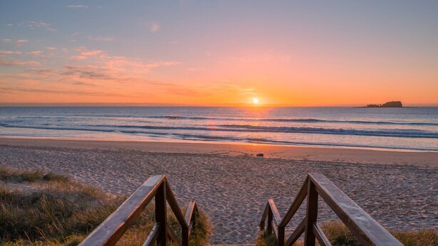 A beach with a wooden walkway leading to the water, a sunset in the background, and a small island visible in the distance. - Makai Hawaiian for direction towards the ocean (Mount Coolum)