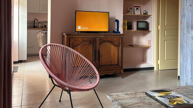 A living room with a television, a wicker chair, a wooden table, and a kitchen in the background. - The Sorbier house, Provence (Maillane)