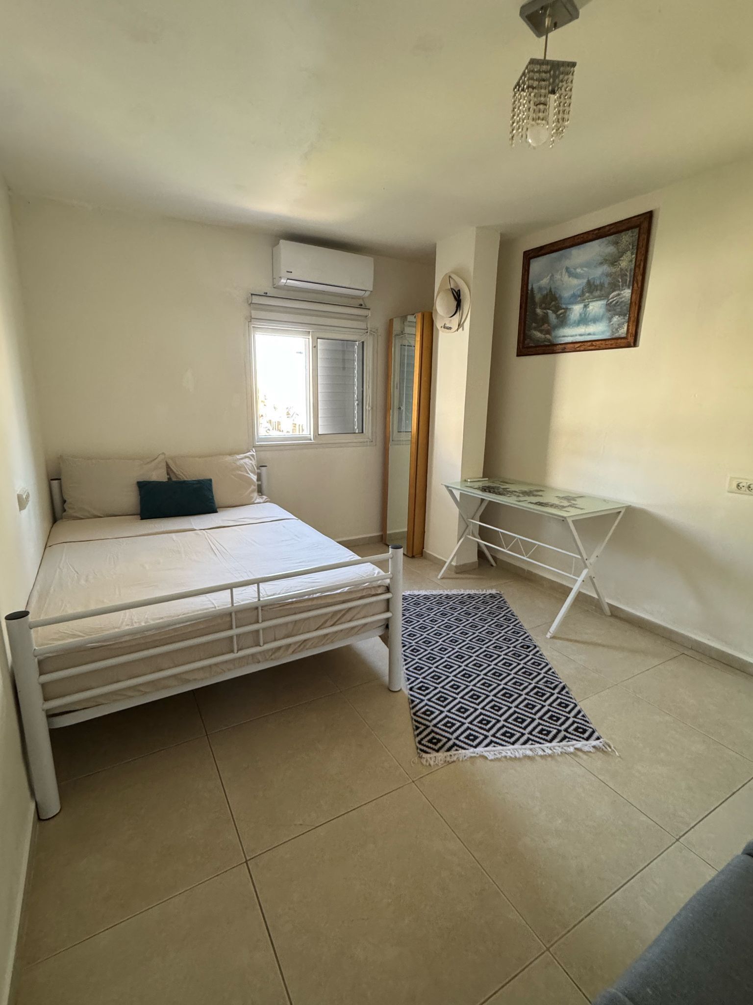 Una camera da letto con un letto, una ventola da terra, una finestra con persiane, un quadro e un piccolo tavolo.