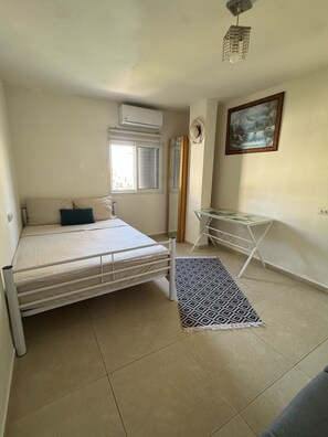 A bedroom with a bed, a standing fan, a window with blinds, a painting, and a small table. - דHuge vue, very central, 10 min. walk from sea, food around, free parking, (Eilat)