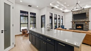 A modern kitchen with a large island, dark cabinetry, and a stone fireplace.