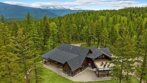 A lodge with a large deck and multiple rooms, surrounded by dense forest and mountains in the background.