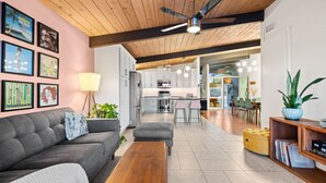 A modern living room with a sofa, ottoman, and coffee table. There is a kitchen in the background with white cabinets and a dining area with chairs. - Golden Hour Bliss: Pool, Hot Tub & Firepit (La Mesa)