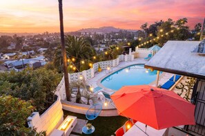 A swimming pool with a view of a residential area, surrounded by palm trees and string lights.