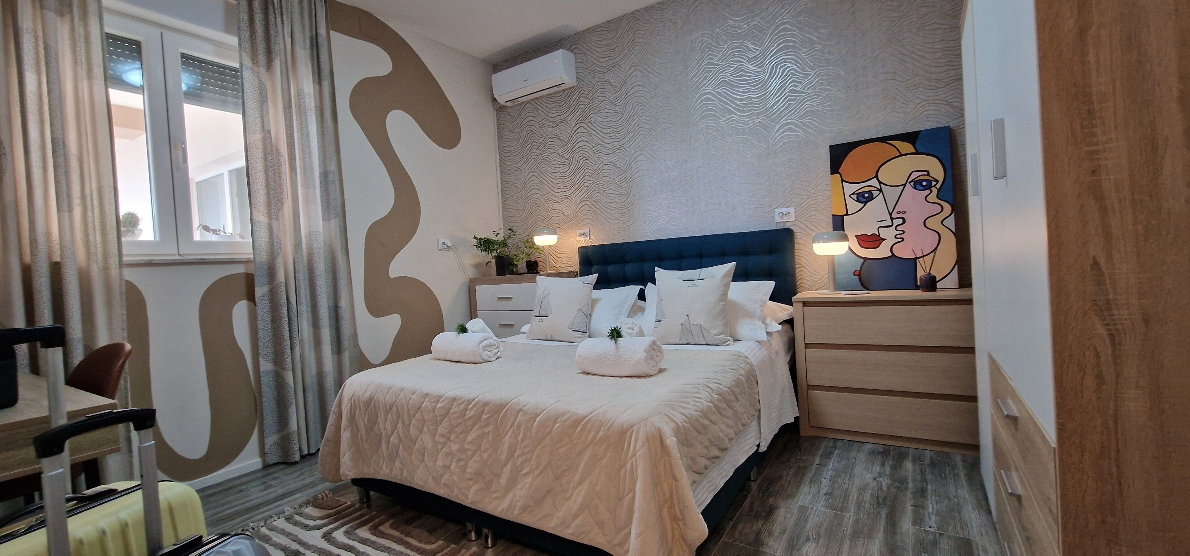 A neatly made bed with a quilted cover, a bedside table with a lamp, and a wall with a large abstract painting.
