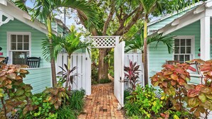 A brick pathway leading to a white gate with a lattice top, surrounded by lush tropical plants and a tree. - ~MARGARITA COTTAGE~ Bask in sunshine and lounge by your pool in paradise. (Key West)