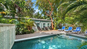 A swimming pool with lounge chairs, surrounded by lush greenery and a small building with a tiled roof.