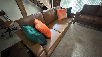 Eine braune Ledersofa mit türkisenen und orangefarbenen Kissen, eine Holztreppe und ein Fenster mit Jalousien.