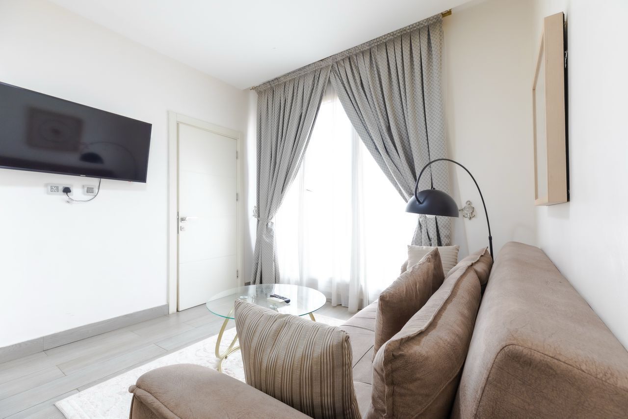A modern living room with a flat-screen TV mounted on the wall, a glass coffee table, a beige sofa, and a floor lamp.