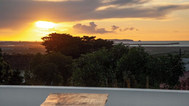Vista del atardecer desde un balcón con piso de madera, árboles y una costa lejana.