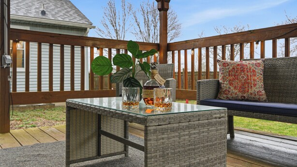 A glass-top table with two glasses and a bottle, a wicker bench with a cushion, and a wooden deck with a fence in the background. - The Bourbon Trail Bungalow Ping Pong/Foosball/Bar/Firepit (Mount Washington)