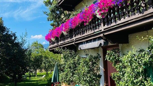 A traditional building with balconies adorned with vibrant flowers, outdoor seating, and a sign reading "Rainhof." - Pension Rainhof (Kitzbuhel)
