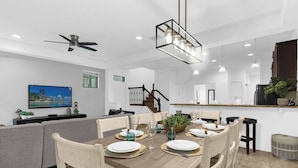A modern dining area with a wooden table set for six, surrounded by white chairs. A ceiling fan and pendant lights are visible above the table. - Dluxx Tranquil 4BR Cozy Home w Full Kitchen 8115 (Davenport)