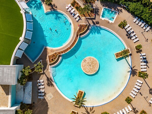 A large, multi-pool resort with a central circular pool, surrounded by sun loungers and palm trees.