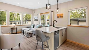 A modern kitchen with a central island, large windows, and a dining area with a sofa and chairs.