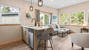 A modern kitchen with a central island, a dining area with a sofa and chairs, and large windows offering views of trees. - SS#8-Kitesurfer-Spring Cove, Terrace Beach, Sauna! (Ucluelet)