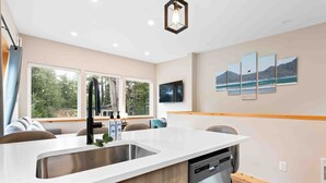 A modern kitchen with a white countertop, stainless steel sink, and a dishwasher. There are large windows providing a view of the outdoors. - SS#8-Kitesurfer-Spring Cove, Terrace Beach, Sauna! (Ucluelet)