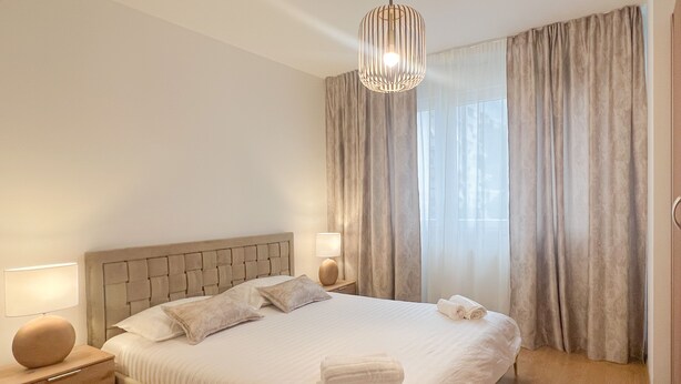 A modern bedroom with a large bed, bedside lamps, a hanging light fixture, and sheer curtains.