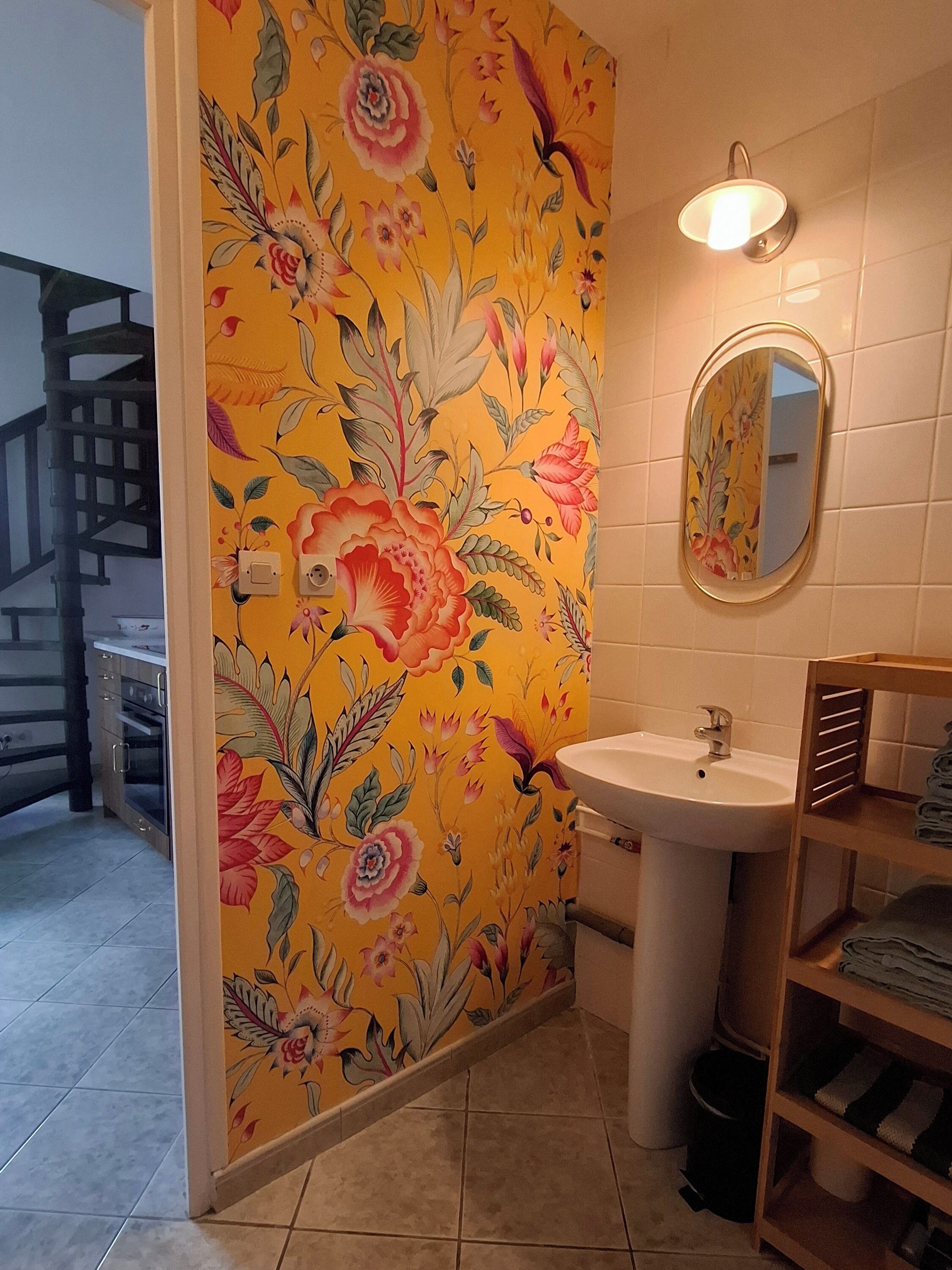 A bathroom with a floral wallpaper, a sink, a mirror, and a wooden shelf with towels.