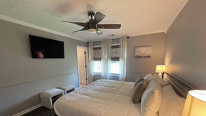 Une chambre d’hôtel avec un grand lit, un ventilateur de plafond, un téléviseur à écran plat fixé au mur, une table de chevet avec une lampe et une fenêtre avec des rideaux.