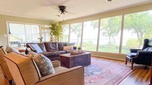 A spacious living room with a large window, a ceiling fan, and a wooden floor. - Lakefront fun for 14+pets w/game room deck sunsets (Whitney)
