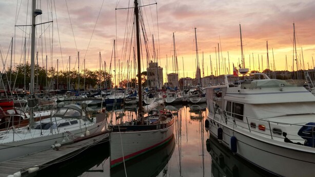 Un port de plaisance où sont amarrés de nombreux bateaux, un ciel dégagé et un coucher de soleil en arrière-plan.