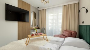 Ein Hotelzimmer mit einem Bett, einem Nachttisch mit Tasse und Blumenvase, einem Sessel, einem an der Wand montierten Fernseher und einem großen Fenster mit Vorhängen.