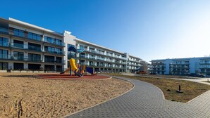 Ein Spielplatz mit Rutschen und Sandkasten, umgeben von einem gepflasterten Weg und einem Gebäude mit mehreren Balkonen.