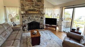 A living room with a stone fireplace, a ceiling fan, a flat-screen TV, and a large window with a view. - Our Shangri La (Berkeley Springs)