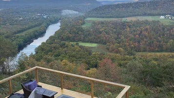 Une terrasse en bois, meublée de mobilier en osier, qui offre une vue sur une rivière et des collines boisées.