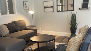 Een woonkamer met een grijze modulaire bank, een ronde salontafel, een getextureerd vloerkleed, een potplant en lijstwerken aan de muur.