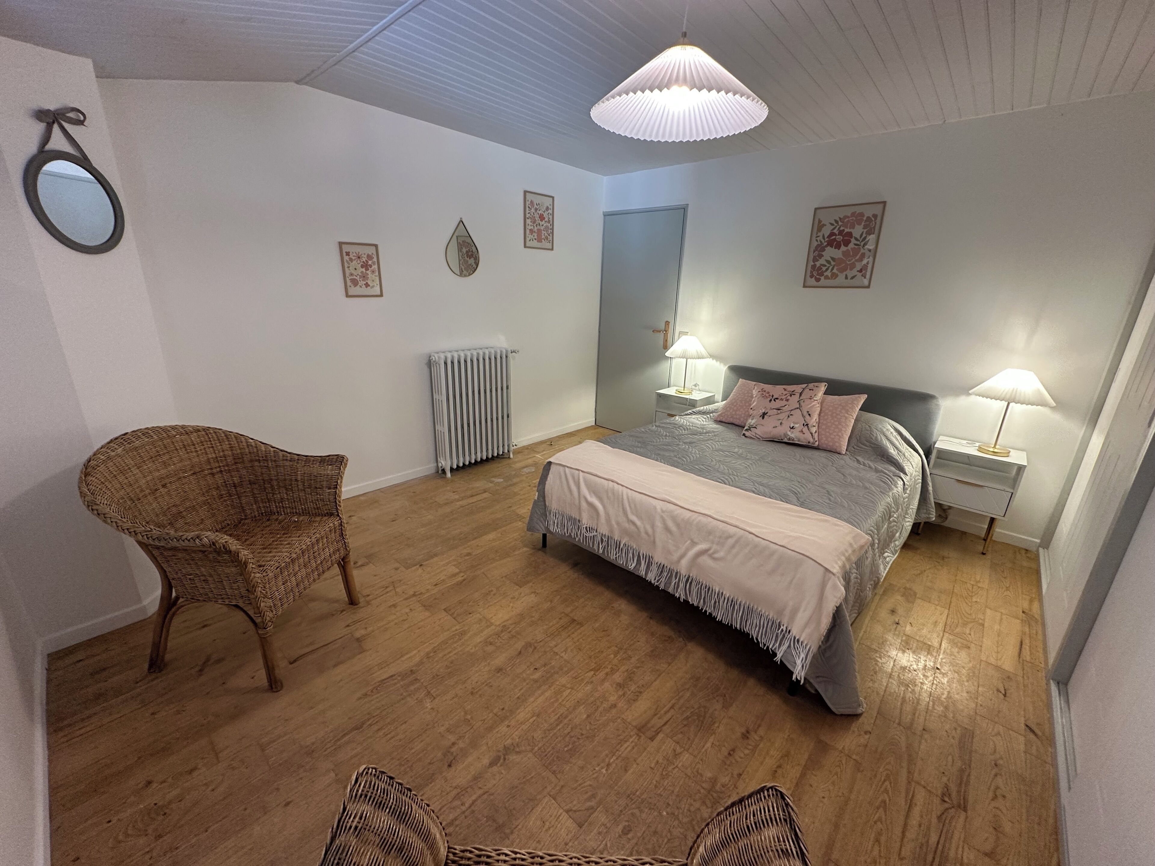 Un dormitorio con cama, dos mesitas de noche con lámparas, una silla de mimbre, radiador y adornos en la pared.