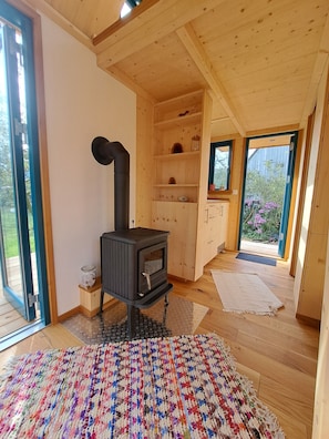 A wooden interior with a black wood stove, a large window, and a colorful woven rug.