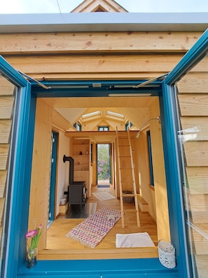 A wooden cabin with a loft bedroom, a fireplace, and a large window.