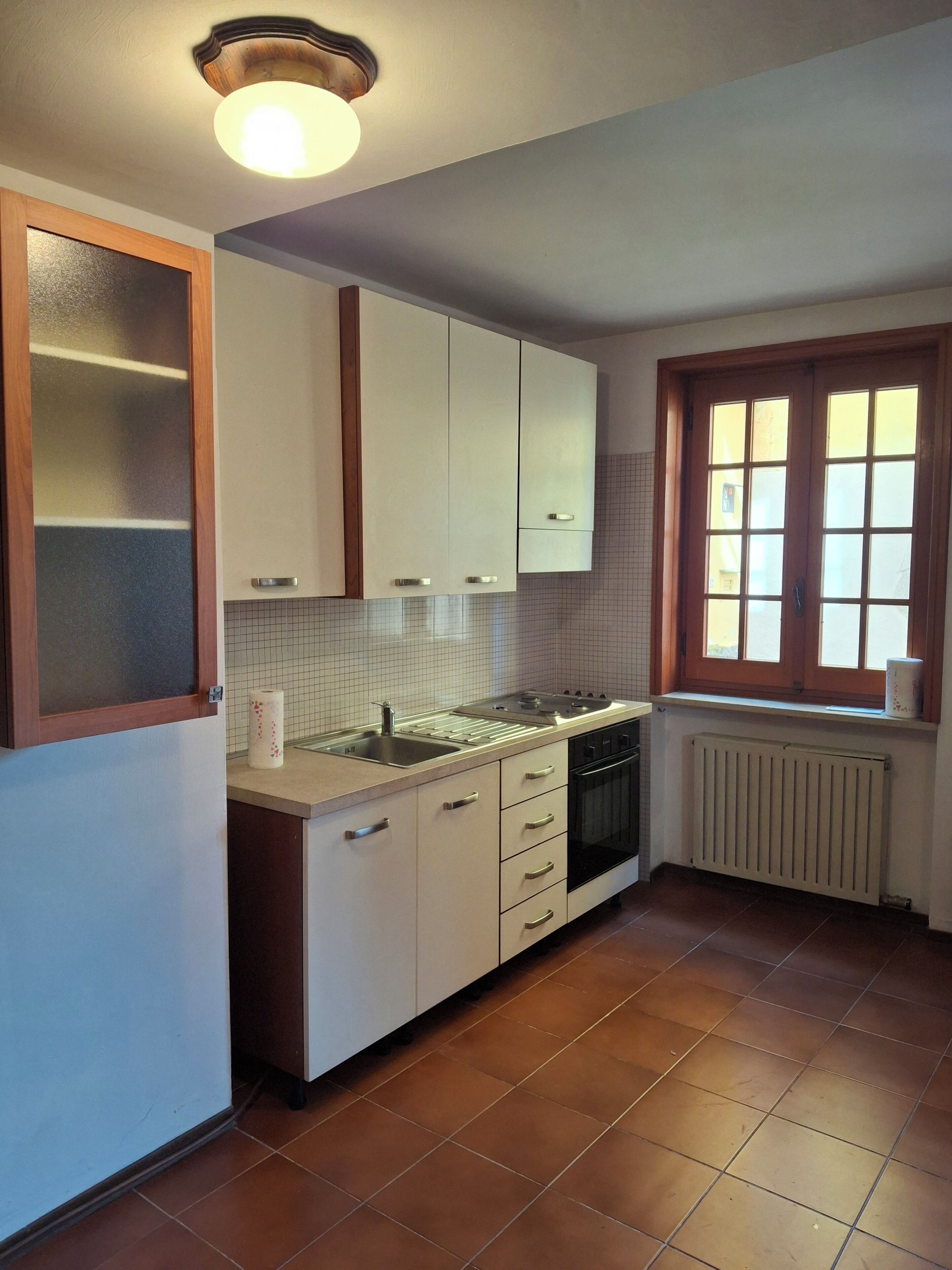 Una cocina con armarios blancos, una ventana con marcos de madera, un radiador y suelo de baldosas.