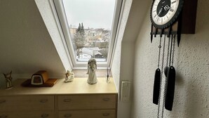 Ein Zimmer mit schrägen Wänden, einem Fenster mit Blick auf eine verschneite Landschaft, einer Wanduhr und einer Kommode mit Schubladen und einer kleinen Figur.