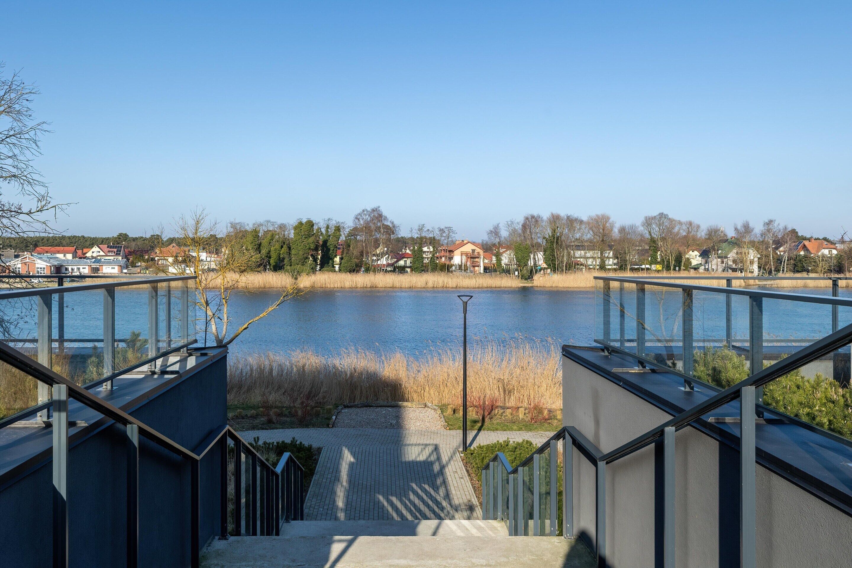Blick von einem modernen Gebäude mit Glasgeländer auf einen ruhigen See, umgeben von Wohngebäuden und Bäumen.