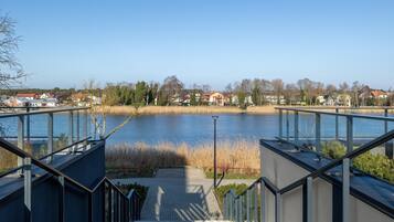 Blick von einem modernen Gebäude mit Glasgeländer auf einen ruhigen See, umgeben von Wohngebäuden und Bäumen.